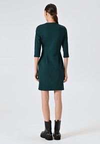 Groene jurk met driekwart mouwen, aansluitende silhouet en een subtiele naaddetail aan de achterkant. Gepaard met zwarte veterschoenen tot de enkel.