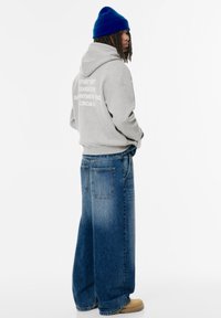 Grå oversized hoodie med vit grafisk text på ryggen, ihopsatt med lösa blå denimbyxor och ljusbruna skor. Blå stickad mössa på huvudet.