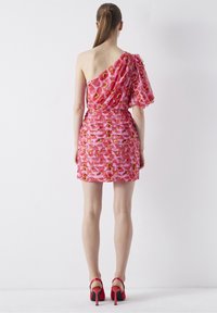 Femme portant une mini-robe rose à fleurs à une épaule avec une manche bouffante et des chaussures à talons hauts rouges, debout devant un fond blanc.