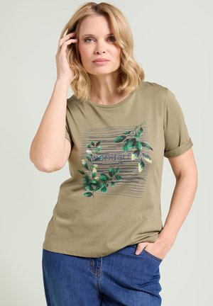 Donna bionda che indossa una maglietta verde oliva con fantasia di foglie verdi e testo, abbinata a jeans blu, posando con una mano vicino al viso.