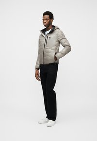 Άνδρας όρθιος που φοράει ανοιχτό γκρι καπιτονέ μπουφάν με κουκούλα, μαύρο παντελόνι, λευκά αθλητικά παπούτσια, με το ένα χέρι στην τσέπη του μπουφάν μπροστά σε μονόχρωμο φόντο.