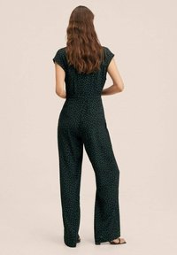 Žena s dlouhými vlnitými vlasy, oblečená v černém overal s malými tyrkysovými puntíky, stojící bosa před jednoduchým béžovým pozadím.