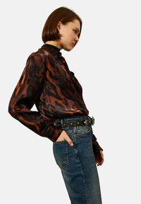 Bluzka w panterkę w ciemnym brązie i czarnym satynowym materiale, z wysokim kołnierzem. Noszona z niebieskimi dżinsami z wysokim stanem i czarnym paskiem z ćwiekami.