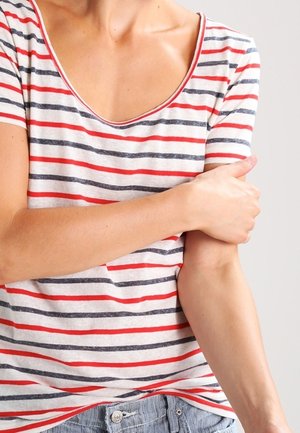 Personne portant une chemise rayée rouge et noire, tenant le haut de son bras gauche avec sa main droite sur un fond neutre.