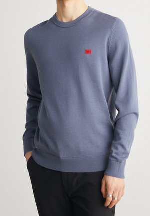 Homme portant un pull bleu clair à col rond avec un petit patch logo rouge sur la poitrine et un pantalon noir, mains partiellement dans les poches.