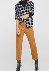 Camicia a flanella a quadri in blu navy e verde, abbinata a pantaloni in velluto a coste color cammello. Il modello indossa stivaletti verdi. Materiali strutturati, vestibilità su misura.