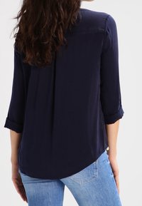 Kvinna med långt brunt hår iförd en marinblå långärmad blus och ljusblå jeans, sedd bakifrån.