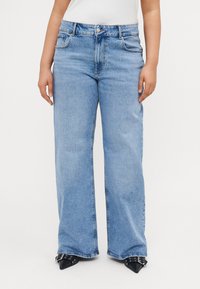 Gaiši zili plataji džinsi, izgatavoti no denim auduma, ar priekšējiem kabatām, pogu aizdari un nodilušu tekstūru pie apakšmalas.