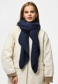 Écharpe côtelée bleu marine en matière douce, drapée élégamment autour du cou, contre une veste beige clair avec des accents marron.