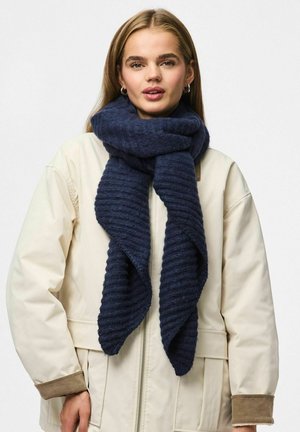 Marinblå ribbade halsduk gjord av mjukt material, draperad elegant runt halsen, mot en ljus beige jacka med bruna detaljer.