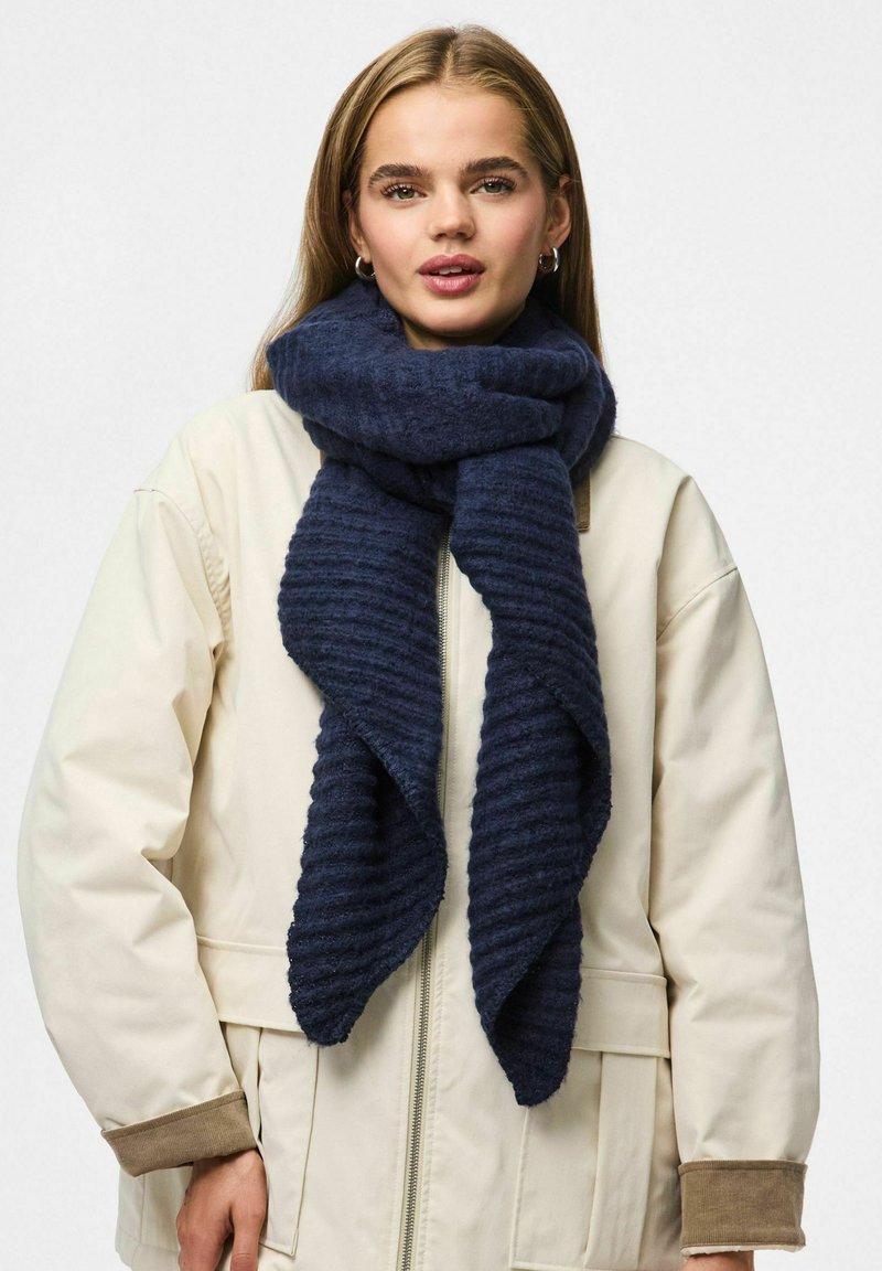 Écharpe côtelée bleu marine en matière douce, drapée élégamment autour du cou, contre une veste beige clair avec des accents marron.