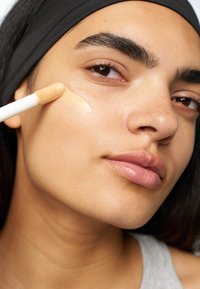 Flytende foundation blir påført huden med en svampapplikator. Huden er glatt, lys brun med fregner, og leppene er naturlig rosa.