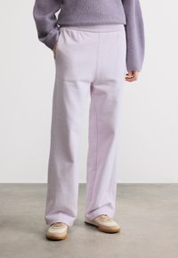 Άτομο που φοράει ανοιχτό μοβ παντελόνι ψηλόμεσο, πλεκτό πουλόβερ μωβ και μπεζ αθλητικά παπούτσια, στέκεται σε  γκρι πάτωμα μπροστά σε λευκό τοίχο.