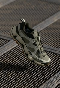 Olive green athletic shoe with mesh and rubber materials, textured surface, unique wave-like design, and lace-up closure with a rubber sole.