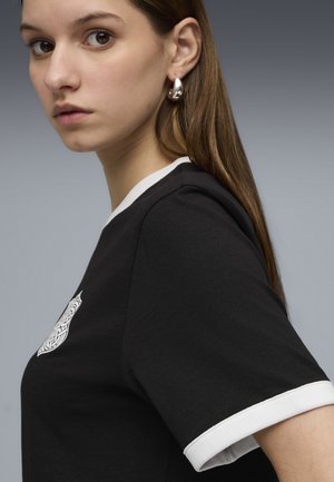 Giovane donna con lunghi capelli castani che indossa orecchini a cerchio d'argento e una maglietta nera con bordi bianchi e un emblema bianco sul petto.