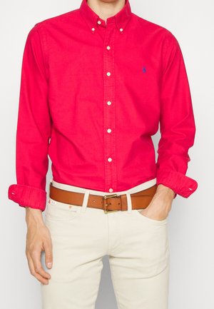 Mężczyzna w jaskrawoczerwonej, zapinanej na guziki koszuli z podwiniętymi rękawami, białych spodniach i brązowym skórzanym pasku z mosiężną klamrą, jedną ręką w kieszeni.