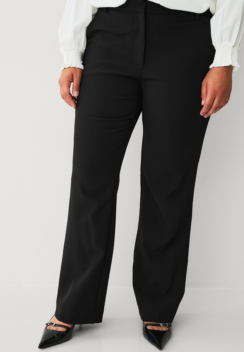 Vrouw draagt zwarte rechte broek met hoge taille, gecombineerd met zwarte pumps met spitse neus en een witte blouse met lange mouwen.