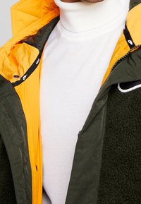 Grön och gul jacka med ett kviltat yttre och en plyschfoder. Vit polotröja under, med en logotyp på jackans dragkedjeområde.