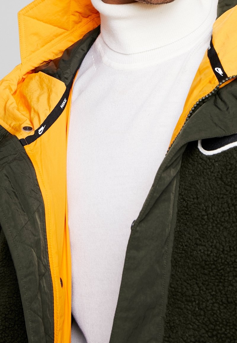 Grön och gul jacka med ett kviltat yttre och en plyschfoder. Vit polotröja under, med en logotyp på jackans dragkedjeområde.