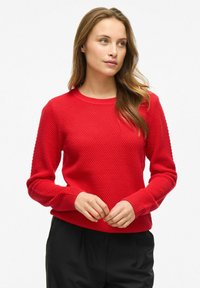 Pull en tricot rouge avec un motif en nid d'abeille texturé, encolure ronde et poignets et taille côtelés, associé à un pantalon noir.