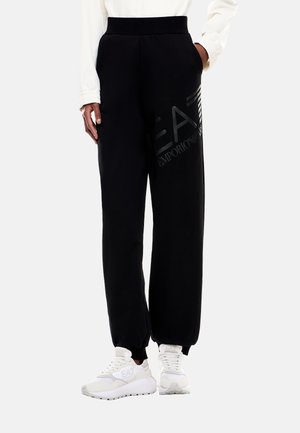 Persona che indossa pantaloni della tuta neri a gamba larga con logo EA7 Emporio Armani sulla coscia e sneakers bianche con logo, mani nelle tasche.