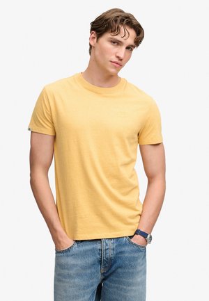 Joven con cabello castaño que lleva una camiseta amarilla de manga corta y vaqueros azules con las manos en los bolsillos contra un fondo liso.