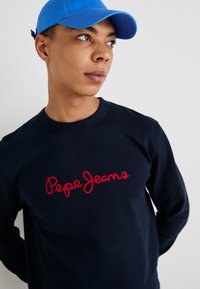 Sudadera navy con el logotipo "Pepe Jeans" bordado en rojo, cuello redondo, tejido texturizado y gorra azul. Diseño simple con acentos mínimos.
