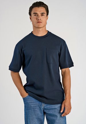 Camiseta de manga corta en un oscuro color marino, hecha de algodón, con cuello redondo y bolsillo en el pecho, combinada con jeans azules. Textura suave.