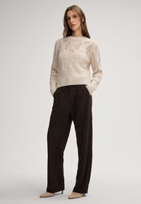 Häkelpullover in Off-White mit offenen Mustern und ribbeligem Saum, kombiniert mit hochtaillierten, schokoladenbraunen weitem Hosen und beigen Absätzen.