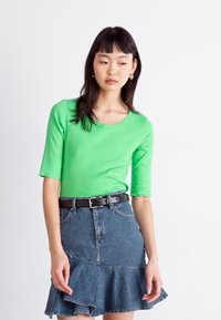 Haut à manches longues vert à côtes associé à une jupe en denim bleu avec un ourlet froncé et une ceinture noire, sur fond uni.
