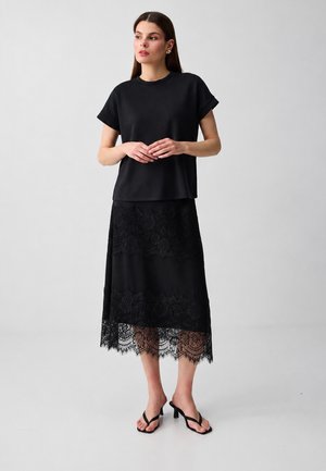 Femme debout, portant un haut noir ample à manches courtes, une jupe midi en dentelle noire et des sandales à talons noires, sur un fond clair uni.