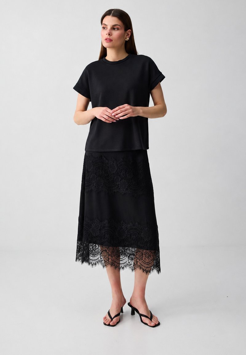 Femme debout, portant un haut noir ample à manches courtes, une jupe midi en dentelle noire et des sandales à talons noires, sur un fond clair uni.