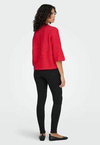 Rdeča zgornja majica z teksturo, kratka, s ¾ rokavi, kombinirana s črnimi ozkimi kavbojkami in črnimi balerinkami. Jasno zadnji pogled, ki prikazuje podrobnosti oblačila.