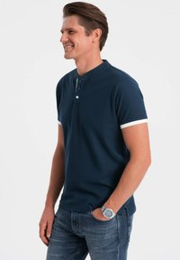 Námorně modré polo tričko s krátkým rukávem, s henley límečkem a bílými akcentními pruhy na rukávech. Materiál vypadá strukturovaně. Nosené s džínovými kalhotami.