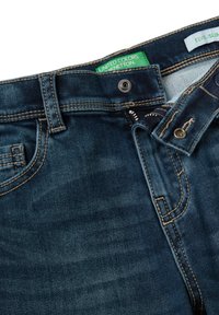 Tmavě modré džíny s úzkým střihem. Obsahují kovový knoflík, zip a kontrastní žluté prošití. Na etiketě je nápis "United Colors of Benetton."