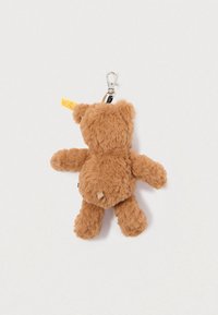 Llave de peluche marrón en forma de oso, con un gancho, que presenta una superficie suave y texturada, orejas redondeadas y una pequeña etiqueta en el lateral.