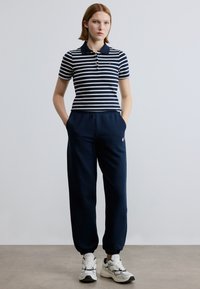 Μπλε ναυτικό ριγέ πόλο με γιακά, συνδυασμένο με μπλε ναυτικό φόρμας και λευκά αθλητικά παπούτσια. Η εμφάνιση έχει άνετη εφαρμογή και casual στυλ.