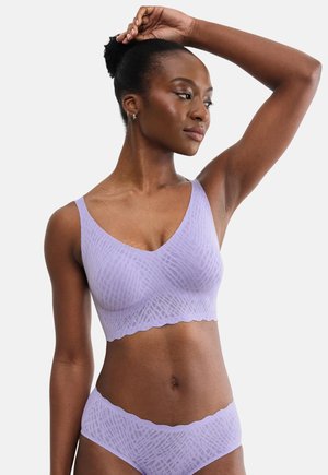 Femme à la peau foncée portant un soutien-gorge et un sous-vêtement violet clair assortis et sans couture, posant avec un bras levé derrière la tête.