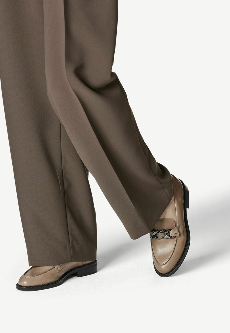 Pantalon marron à jambes larges associé à des mocassins en cuir beige avec un détail de chaîne noire sur des semelles plates.