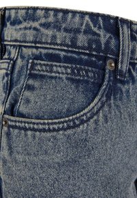Denim teksad on tume sinine pesu, nähtavad õmblused, eesmine tasku ja metallne popp. Kanga välimus on veidi tekstureeritud.