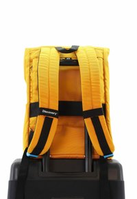 Mochila amarilla con correas acolchadas unida a una maleta negra, con una textura de tela y un logo de Discovery en una de las correas.