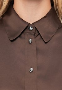 Καφέ πουκάμισο με κλασικό γιακά, έξι λαμπερά κουμπιά στο μπροστινό μέρος και απαλό, ελαφρώς γυαλιστερό ύφασμα.
