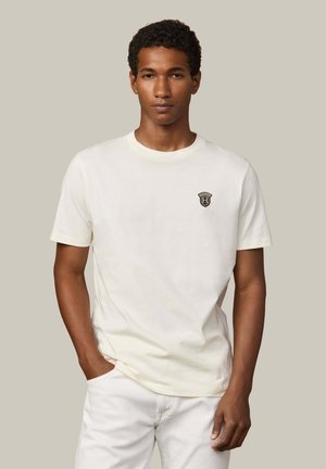 Crème kleurige t-shirt met korte mouwen van zachte katoen, met een klein donker logo op de linkerkant van de borst. Ronde halslijn en een relaxte pasvorm.