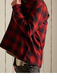 Chaqueta de forro polar a cuadros rojos y negros con cierre de botones, diseño con cuello y tela texturizada, usada sobre una camisa de color claro.