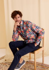 Kleurvolle bloemen overhemd met knopen, met rode en gele bloemen op een marineblauwe achtergrond, gecombineerd met donkere pantalons en grijze instapschoenen.