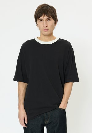 Čierne nadmerné tričko s bielym rebrovaným výstrihom, s krátkymi rukávmi a voľným strihom. Nosené s tmavými denimovými džínsami.