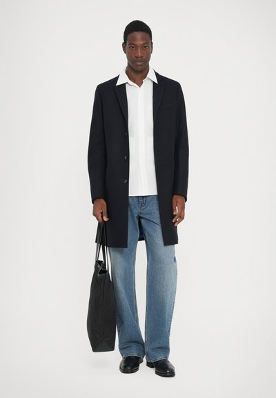 Cappotto navy con un design su misura, camicia bianca, jeans a gamba larga azzurri chiari e una borsa tote nera. Tessuto liscio, linee pulite e accenti minimal.