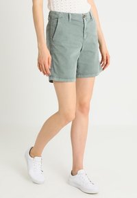 Shorts en coton vert clair à coupe décontractée, dotés de poches latérales, bords ourlés et fermeture par bouton. Portés avec des baskets blanches.