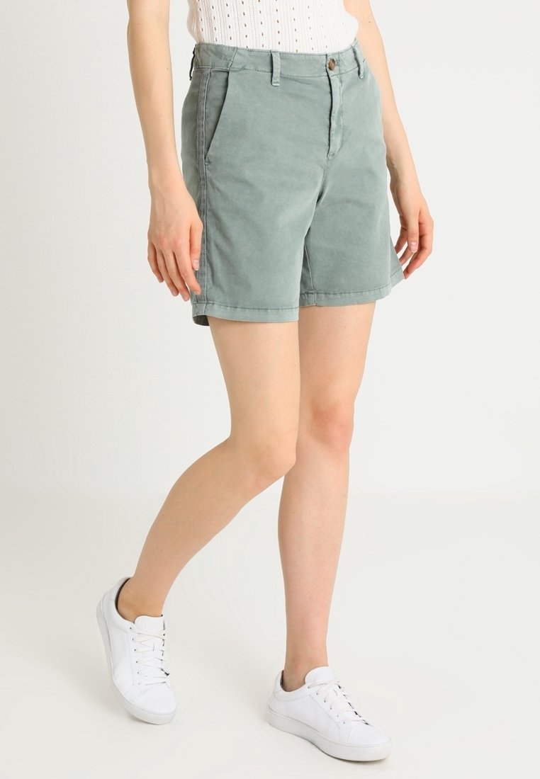 Shorts en coton vert clair à coupe décontractée, dotés de poches latérales, bords ourlés et fermeture par bouton. Portés avec des baskets blanches.