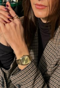 Gold wristwatch with a square dial and a green face, worn on a slender wrist. Long brown hair and a houndstooth-patterned blazer are visible.